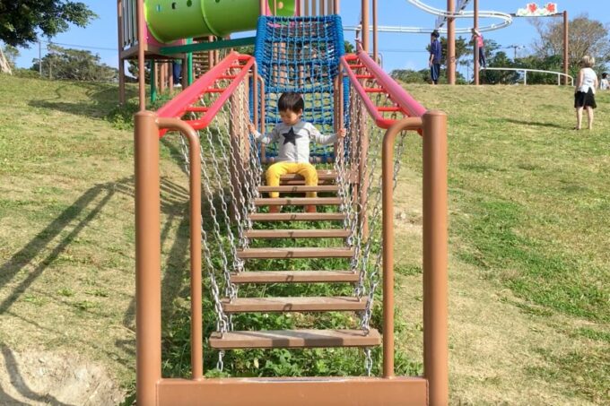 南城市「グスクロード公園」のアスレチック遊具をブランコのように楽しむ幼いお子サマー